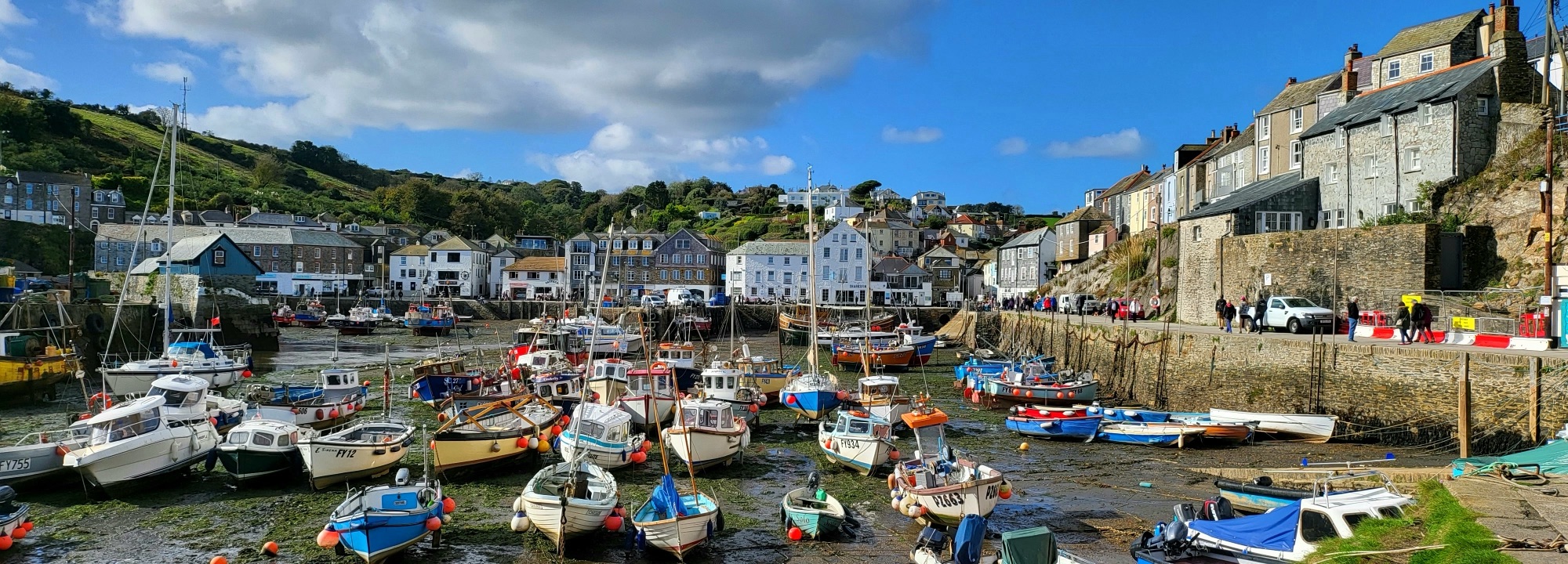 A vibrant harbour scene in Mevagissey, Cornwall, with numerous fishing boats moored in the shallow waters during low tide. The boats, painted in bright colours with red, blue, and white hulls, rest on the muddy seabed, their reflections faintly visible. The harbour is lined with historic stone and pastel-coloured buildings, nestled against a lush green hillside. A stone wall runs along the waterfront, where people stroll and explore the charming fishing village. The sky is a mix of blue and white, with scattered clouds casting soft shadows over the scene.