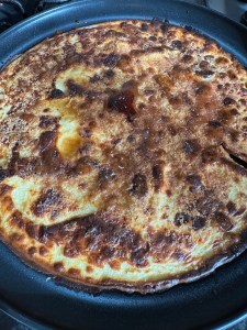 A gluten-free pancake in a black frying pan with a crispy, caramelised surface. The pancake has an uneven golden-brown colour, with some darker, almost burnt spots. There are patches of melted sugar, giving it a glossy appearance in some areas. The edges are slightly curled and crisp, and the texture appears soft in the lighter areas.