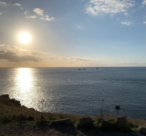 A stunning sunset over the Atlantic Ocean at Land’s End, Cornwall, with the sun casting a golden glow across the water. The sky is a blend of soft blues and warm oranges, with scattered clouds adding depth. In the distance, a lighthouse stands on a rocky outcrop surrounded by small islands. The foreground features a rugged, grassy cliff with scattered stones, overlooking the vast, calm sea.
