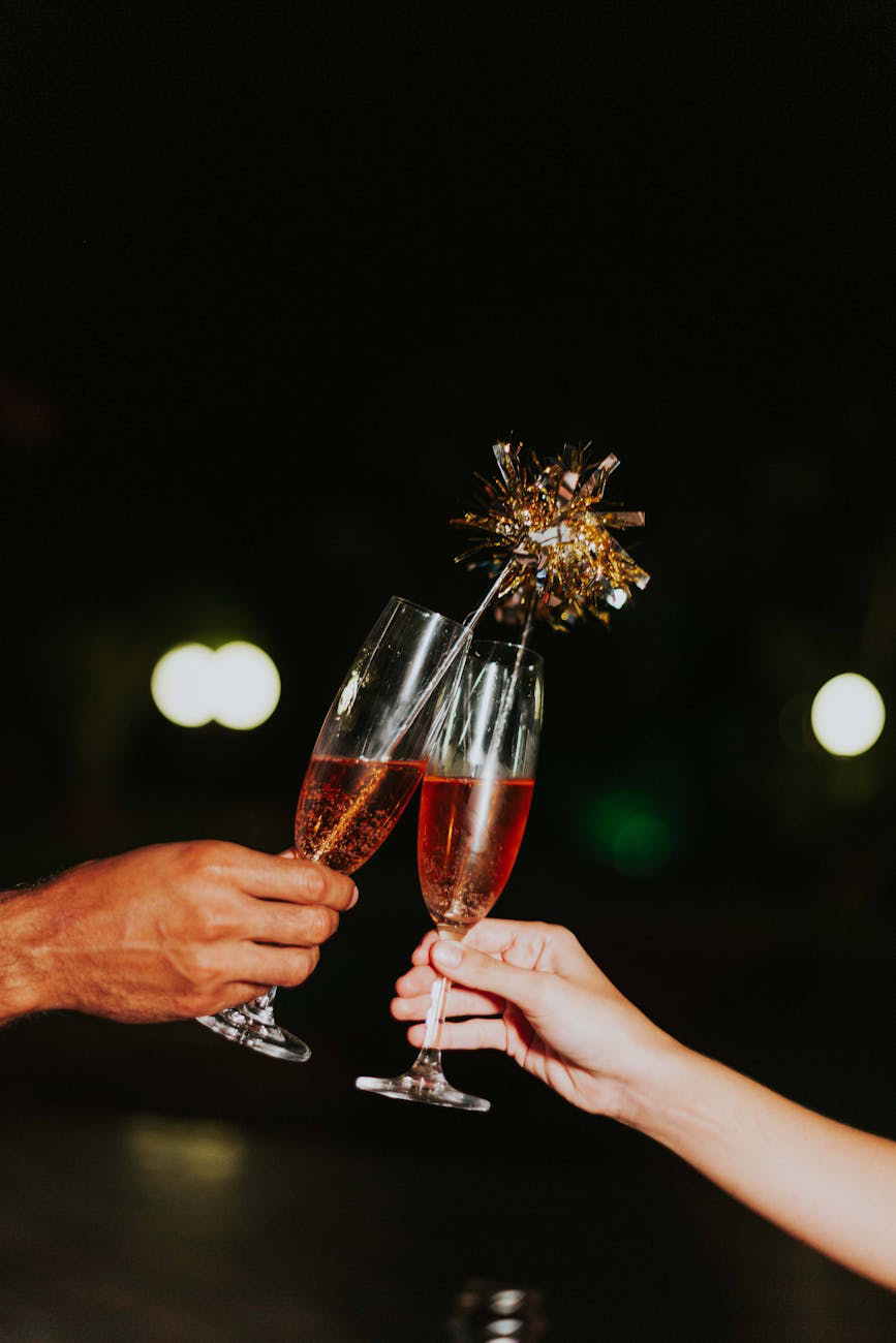 A close-up photograph of two people toasting with champagne flutes filled with sparkling rosé wine. One hand appears darker in complexion, while the other is lighter, suggesting diversity. The glasses clink together, with one decorated with a gold tinsel-like ornament. The background is blurred with glowing bokeh lights, creating a festive and celebratory atmosphere.