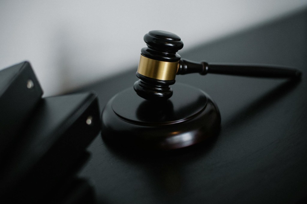A close-up photograph of a judge’s gavel resting on a wooden sound block. The gavel is dark wood with a gold band around its head. The image has a moody, dimly lit atmosphere with a blurred background, emphasizing the gavel as the focal point. To the left, there are black binders with silver rivets, suggesting a legal or courtroom setting.