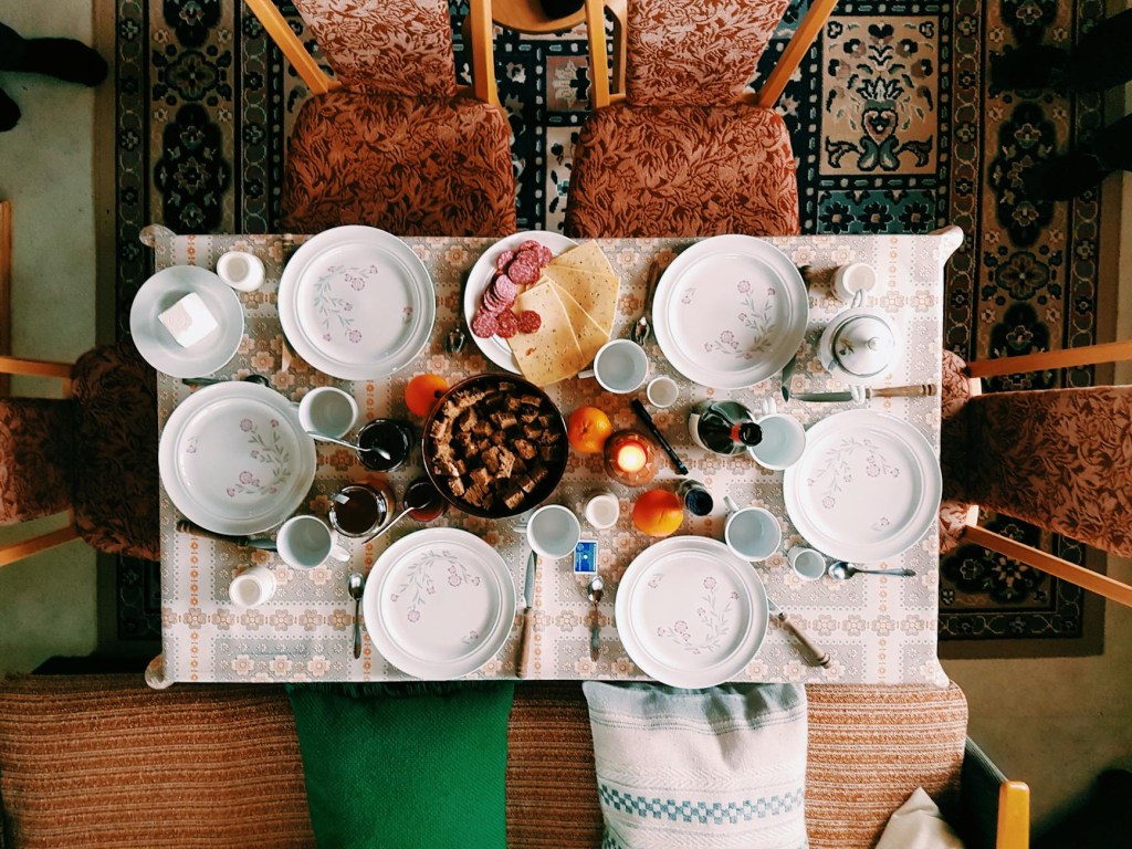 A top-down view of a dining table set for a meal, surrounded by six ornate wooden chairs with patterned cushions. The table is covered with a vintage-style tablecloth and set with floral-patterned plates, teacups, and cutlery. In the center, there is a bowl of cubed bread or croutons, alongside a plate of sliced salami and cheese. There are also oranges, a lit candle, a teapot, a small dish of sugar cubes, and several glasses of tea. A green blanket and a decorative cushion are draped over a sofa at one end of the table. The surrounding floor is covered with a richly patterned rug, and dark shoes are visible at the edges of the frame, suggesting people nearby.