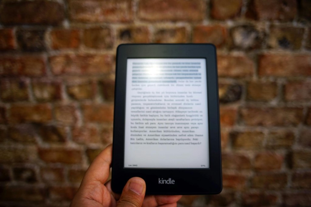 Photo of a man’s hand holding a black kindle in front on a brick wall.