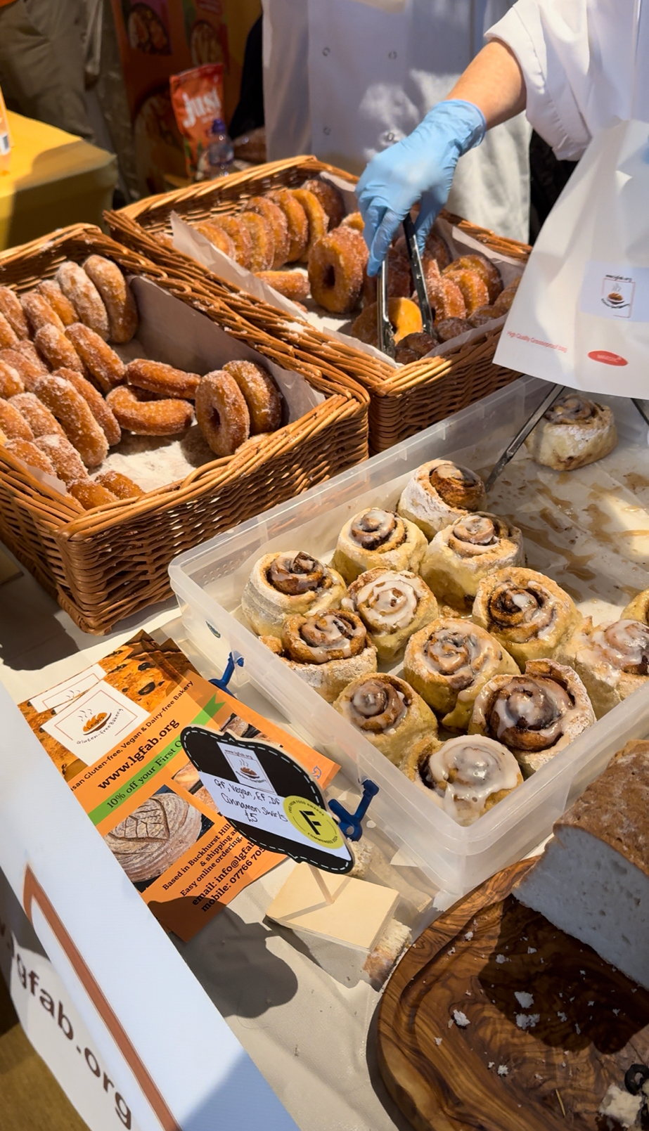 A display of gluten-free baked goods at a food event, featuring sugar-coated doughnuts in wicker baskets and cinnamon rolls with icing in a plastic container. A vendor wearing blue gloves is using tongs to pick up a doughnut.