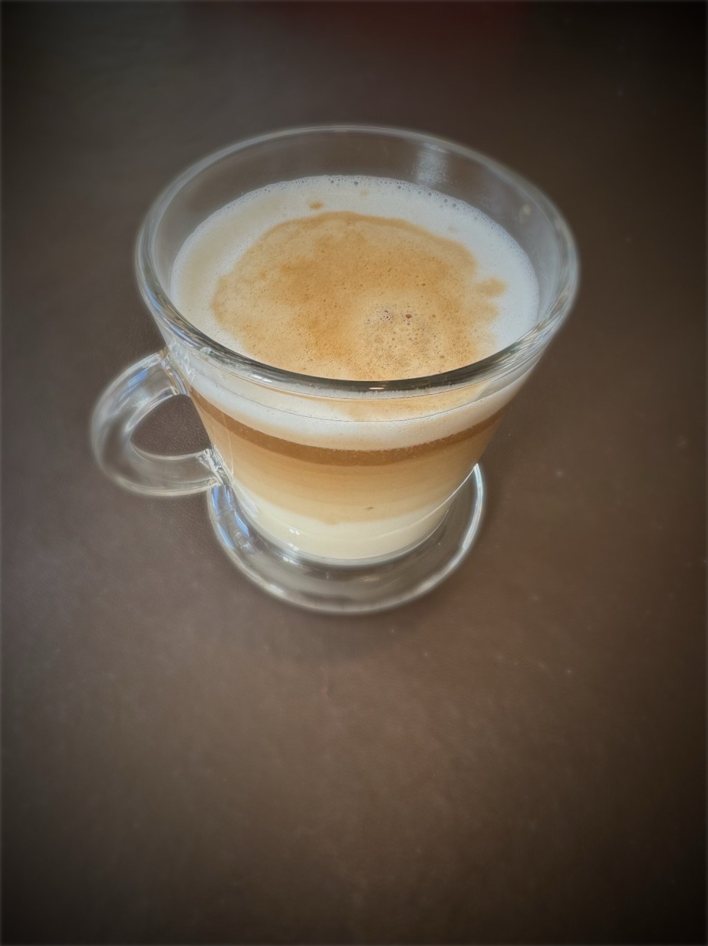 A glass mug with a handle, filled with a layered latte macchiato. The drink has distinct layers of steamed milk, espresso, and frothy milk foam on top. The mug sits on a dark brown surface, with soft lighting highlighting the textures and colours of the coffee.