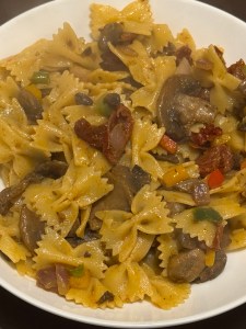 A white bowl filled with gluten-free bowtie pasta mixed with sliced mushrooms, diced red onion, red and yellow peppers, and strips of sun-dried tomato. The pasta is coated in a light, spiced oil-based sauce, giving it a warm, golden colour.
