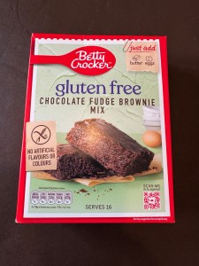 Box of Betty Crocker Gluten Free Chocolate Fudge Brownie Mix on a dark surface, showing the front packaging with an image of brownies and the label highlighting “No artificial flavours or colours.”