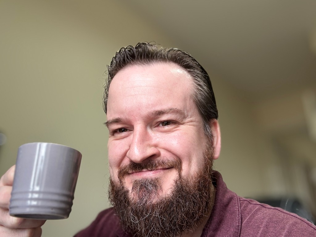 A bearded man with slicked-back dark hair and a maroon polo shirt smiling warmly while holding a grey mug of coffee towards the camera.