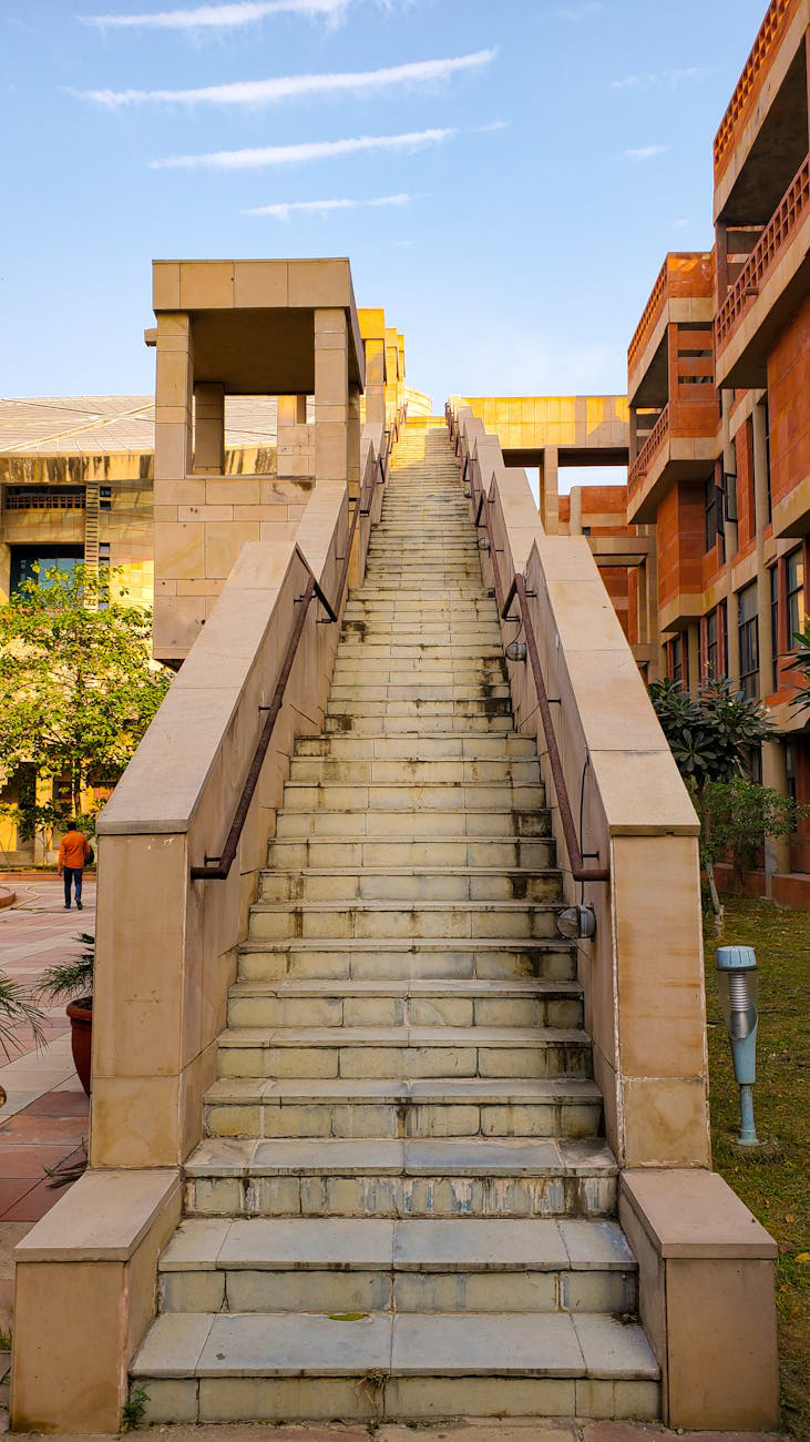 A long, wide outdoor staircase made of light-colored stone or tile ascends upwards. The staircase has railings on both sides made of dark metal bars supported by the stone structure. The stairs lead towards the top of a gentle incline, with buildings visible on either side under a partly cloudy blue sky.