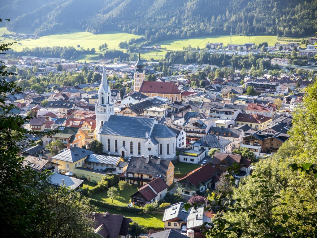 A medium-distance, high-angle photograph shows a town with a prominent white church featuring a tall, pointed steeple and a light blue roof at its center. The town is densely packed with houses featuring red roofs, surrounded by green trees and foliage. The town is located in a valley with rolling green hills and mountains in the background under a bright, sunny sky. The image is taken from an elevated position, possibly a hillside, with trees visible in the foreground. Choose the option that best suits your needs and the context in which the image will be used.