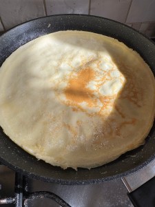 A close-up, overhead shot of a pale, crepe-style pancake in a dark speckled pan on a stovetop. Bright sunlight casts a shadow across the top of the pancake as it turns golden in the middle.