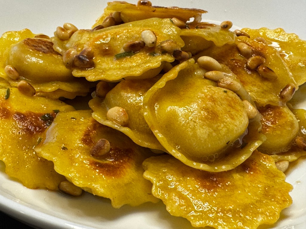 Close-up of gluten free prosciutto and mozzarella girasoli tossed in browned butter with toasted pine nuts on a white plate.