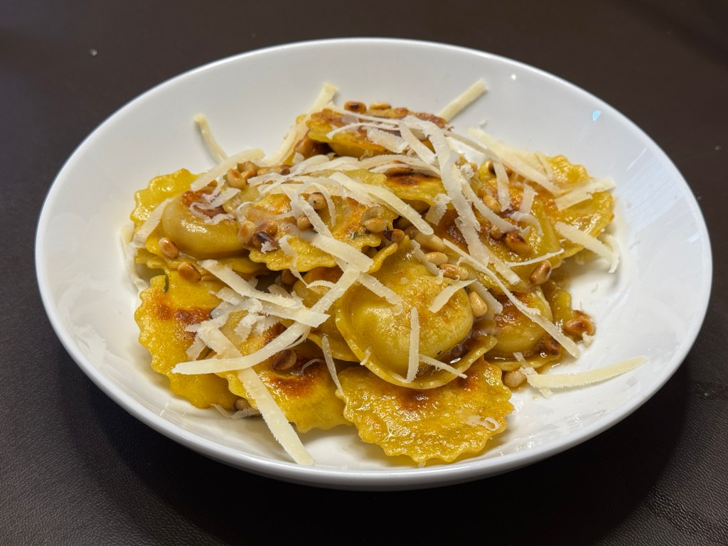 Gluten free prosciutto and mozzarella girasoli tossed in browned butter with toasted pine nuts and topped with Parmigiano Reggiano shavings on a white plate.